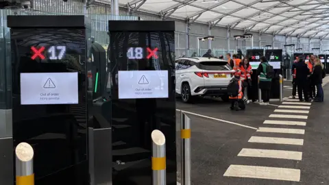 Two finger printing machines in the foreground of a hanger at Eurotunnel in Folkestone with cars and people in the background.