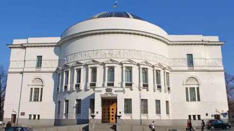 The World Monuments Fund A rectangular white stone building with a curved feature at the front and a dome shaped roof. There are 17 windows across the curved part and a further 4 windows on the flat front of the building. There are steps up to a double wooden door.