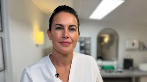 Emma Baugh/BBC Danielle Black is smiling at the camera while wearing a white v-neck top. She has dark hair that has been tied back from her face. Behind her is a mirror and a desk. She appears to be in her practitioner room.