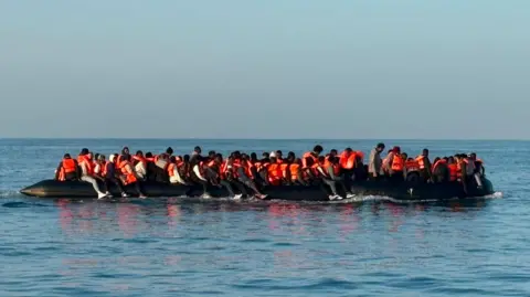 French lifeboat A large black inflatable dinghy carrying dozens of people wearing orange life jackets travels across calm sea water. People are seated along both sides of the vessel and across its centre.