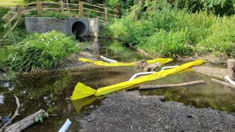 Derbyshire County Council Area at edge of pond where a large concrete pipe opening surrounded by greenery is partly blocked off by floating yellow barriers