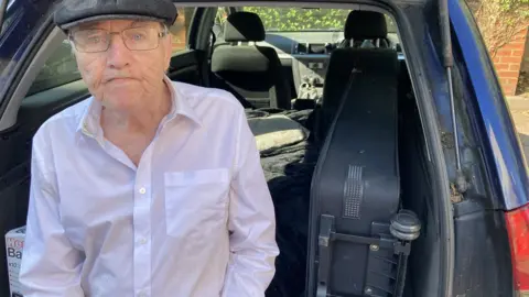 James Grant/BBC Man in flat cap with glasses sits in the boot of a car