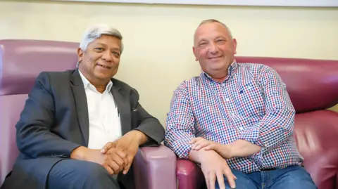 A man, wearing a checked shirt, and another man, wearing a black suit and white shirt. Sat in two purple chairs and smiling at the camera.