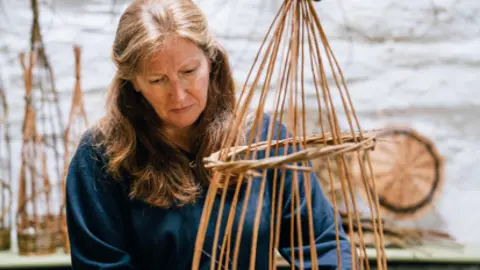 Jo Entwistle Jo standing by a bench with the beginning of a basket being made. She is weaving willow through upright sticks which meet at a point at the top. She is wearing a blue top and jeans and has long brown hair