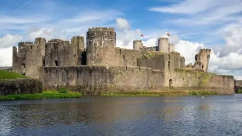 Getty Images Caerphilly Castle with a blue sky and clouds overhead, overlooking a moat. Two Welsh flags can be seen flying from the building.