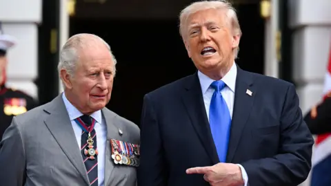 King Charles standing next to US President Donald Trump. who is speaking and pointing towards the monarch.