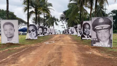 Moses Kollie Garzeawu / BBC A dirt road leading to the sea with black and white photos of those killed on either side, attached to palm trees