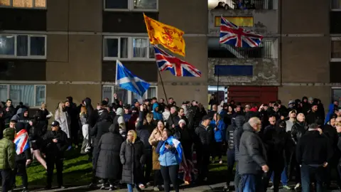 Dozens of people, some of them waving Scottish flags, gather outside a block of flats