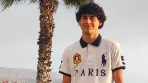 GoFundMe A teenage boy with dark curly hair and wearing a white t-shirt. He is standing at a beach. There is a palm tree behind him.