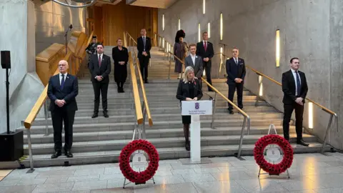 Members of Scottish Parliament dressed in formal attire stand on a staircase in Holyrood in silence paying tribute as a women stands at a podium. 