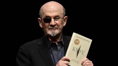 Getty Images Salman Rushdie wearing a black jacket and black shirt, holding up his book Knife. He is wearing glasses with the right lens blacked out.