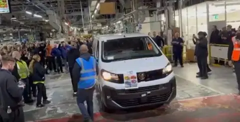Contributed A casual photo taken inside the Vauxhall plant on its last day, showing staff gathered around on the factory floor with a white van in the centre. Some people are taking photos and a few have orange, blue or yellow high vis vests on.