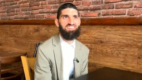 Jody McIntyre is sitting at a table and wearing a light-coloured suit and white shirt