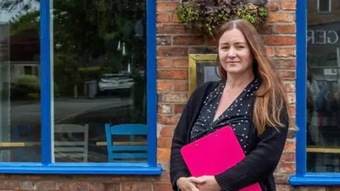 Kelly Woodnutt stood in front of her restaurant St Martha. She is wearing a dark polka dot dress and holding a pink clipboard. Her restaurant is blue.