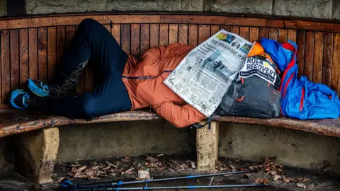 Andrew Hendry / Montane Winter Spine Race A race, wearing blue leggings and an orange jacket, lying on a bench with a rucksack for a pillow and a newspaper over his face