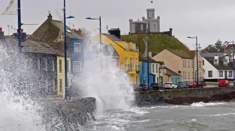 Big waves crashing against the sea wall in Donaghadee. There are a row of colourful houses behind and a moat on top of a hill to the right of the picture.