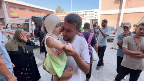 Zeina's head is bandaged and she is carried by her father who is crying, surrounded by people 