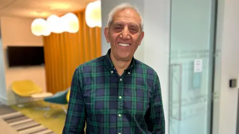 Hardyal Dhindsa smiles in the corridor of Derby City Council's offices in a green chequered shirt 