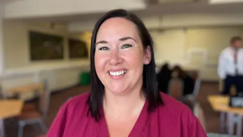 Claire smiles at the camera. She has shoulder length dark hair and is wearing a maroon top.