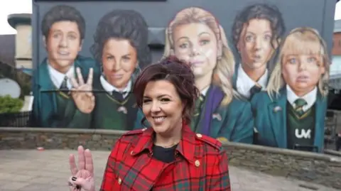 PA Media Derry Girls creator Lisa McGee, in red and green check jacket and holding up three fingers on her right hand, stands in front of the Derry Girls mural. The mural depicts the characters from her hit comedy show.
