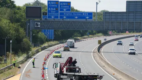 Eddie Mitchell Police and recovery vehicles are in a coned-off section of the motorway. A flatbed recovery truck is parked in the closed lanes while two highway workers in hi-vis clothing stand beside the hard shoulder. Overhead blue motorway signs point to Crawley, East Grinstead, Brighton and Horsham.