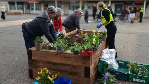 A group of people working together around a large wooden raised planter in an open square, adding soil and arranging colourful flowers. Gardening tools, potted plants and trays are scattered around the planter. In the background, more people are visible near a row of brick buildings with shopfronts and upper‑floor flats. The main workers are three women, and police officer in a high-vis jacket with her blonde hair tied back. 