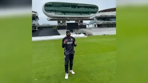 Moshfique Ahmed/Blind Cricket England and Wales Moshfique Ahmed, a man with a black beard and wearing a black t-shirt and baseball camp and a pair of sunglasses, stands on a cricket pitch giving a thumbs up to the camera.