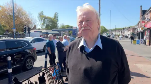 A white man with white hair and a white moustache stands in a shopping street. He is wearing a blue shirt with a black jumper over the top and has his arms crossed behind his back. He is looking slightly to the right as he looks towards the camera, as he is speaking to the reporter who is behind the camera.