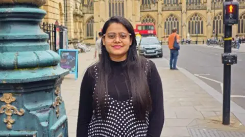 Bhoomi Chauhan missed her flight to London due arriving late at Ahmedabad Airport. Bhumi is posing for a photo next to the backdrop of a city centre landmark, next to a navy lamppost and a pelican crossing