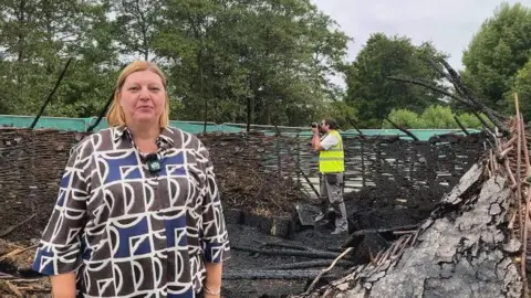 Katherine Ganczakowski/BBC Jacqueline Mooney, with shoulder-length hair parted in the middle and wearing a geometric brown, blue and black shirt, stands in the centre of the charred roundhouse remains. There is a photographer in a high-vis jacket taking photos behind her.