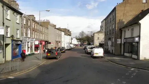 GEOGRAPH / David Dixon Stramondgate in Kendal. It is a town high street with shops and office buildings on each side of the road. There are vans and cars park in the street. The terraced buildings are older in style.
