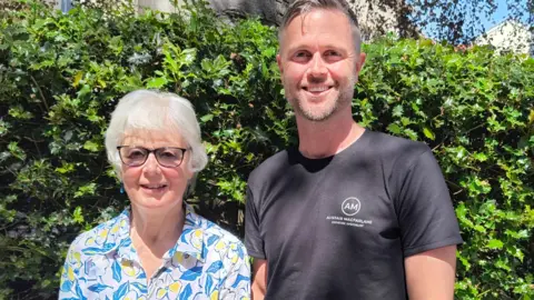 Alastair and his mum Anne are pictured standing in front of a green bush. They are both smiling at the camera. Anne has short white hair and is wearing a white shirt with a blue leaf and yellow flower pattern. She is also wearing black-rimmed glasses. Alastair has short greying hair and stubble. He is wearing a black T-shirt with a white logo.