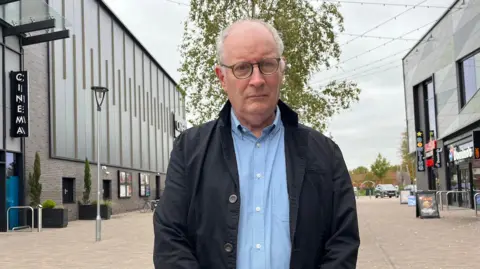 BBC Councillor Tim Mander is looking down the lens with a neutral expression. He;s wearing a blue shirt and dark overcoat. He is stood in the centre of a street, with a cinema on one side and takeaways on the other.