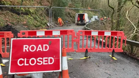 A road closed sign, ahead of safety fencing and workmen beginning preparations for roadworks in the background