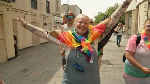 A woman smiling and waving her hands, wearing a pride flag around her neck