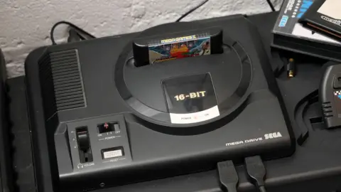 Shaun Whitmore/BBC A black Sega Mega Drive is shown on a black table, with a game cartridge inserted into the machine.