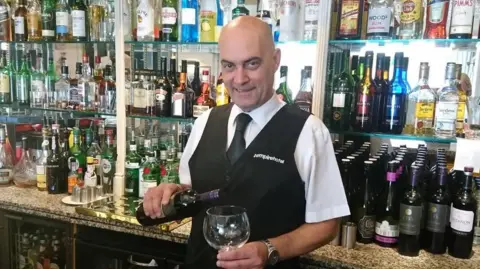 Empire Hotel Jean Paul who is wearing a white shirt, black tie and black waistcoat, pouring a drink behind the bar. There are bottles of alcohol behind him, mainly spirits and red wine.