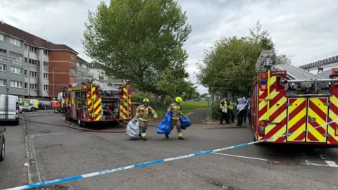 Emergency services at the scene of a chemical incident in Glasgow. Firemen carrying bags are seen walking away from the scene, while two fire engines are parked near blocks of flats. Police vans and officers wearing high-vis vests can also be seen.