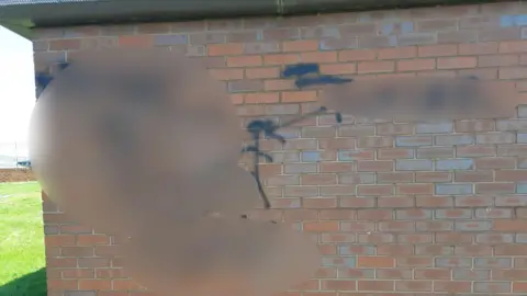 Supplied The red brick wall of the single-storey toilet building has black graffiti on it, a majority of which is blurred out. A stick figure in the centre is the only area which has not been blurred. There is a grassy field and a fence on the left.