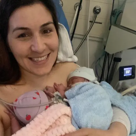 Lowri Price Lowri Price holds her baby twins in a hospital neonatal care unit. One infant wrapped in a pink blanket and another in a light blue blanket.