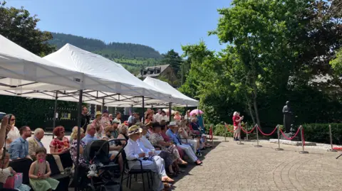 A white marquee with crowds of people sat under it. Acriss from them is a red rope and behind that is the grey statue. It is a sunny day and there are green trees and hills in the background.