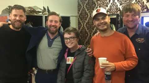 Chloe Russell A woman wearing a black padded jacket and t-shirt smiles into the camera. She is surrounded by the Kaiser Chiefs band who are also smiling.