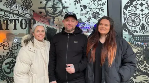 Alice Cullinane/BBC Two women and a man are standing in front of the entrance of a tattoo shop that has patterned skulls and a reflective print over the windows.