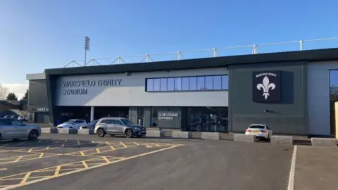 Wakefield Trinity rugby league ground's East Stand at Belle Vue, with blue skies above and a car park in front