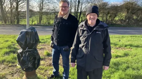 Two men standing next to the stink pipe stump, covered with a black bin liner