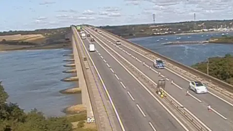 National Highways Four-lane bridge with narrow central reservation. There are cars and vans with gaps between them, There are pontoon supports visible on the waterline. There hedges on both sides of the bridge in the foreground, while fields and buildings can be seen in the background.