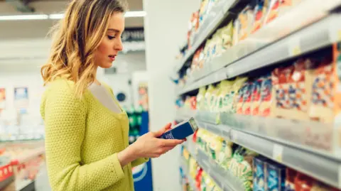 A woman wearing a yellow cardigan looking at an item she has just picked off a supermarket shelf