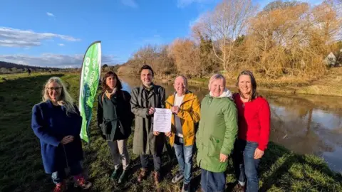 Lewes District Council Six people wearing jackets and jumpers, stood at the side of a river in the sun. Two people in the centre are holding up a document to the camera.