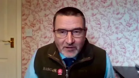 Martin Parker, head of firearms at BASC, is man with short dark hair, glasses, and a beard. He is sitting indoors in front of a floral-patterned wallpapered wall. He is wearing a blue shirt, a dark vest with the "BASC" logo embroidered on it, and a tie.