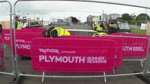 Organisers setting up the event. Crowd control barriers are lined up with the Summer Sessions logs. Workers dressed in hi-vis are on the site. The stage can be seen in the background.